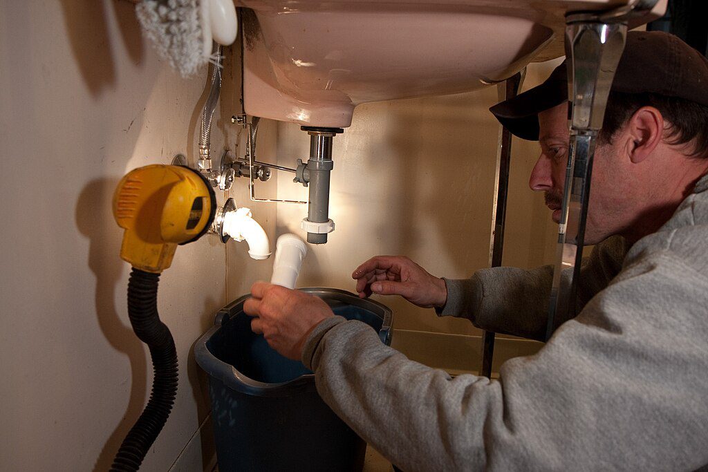Professional plumber doing plumbing service under a bathroom sink, inspecting and repairing drain pipes for proper water flow and leak prevention.