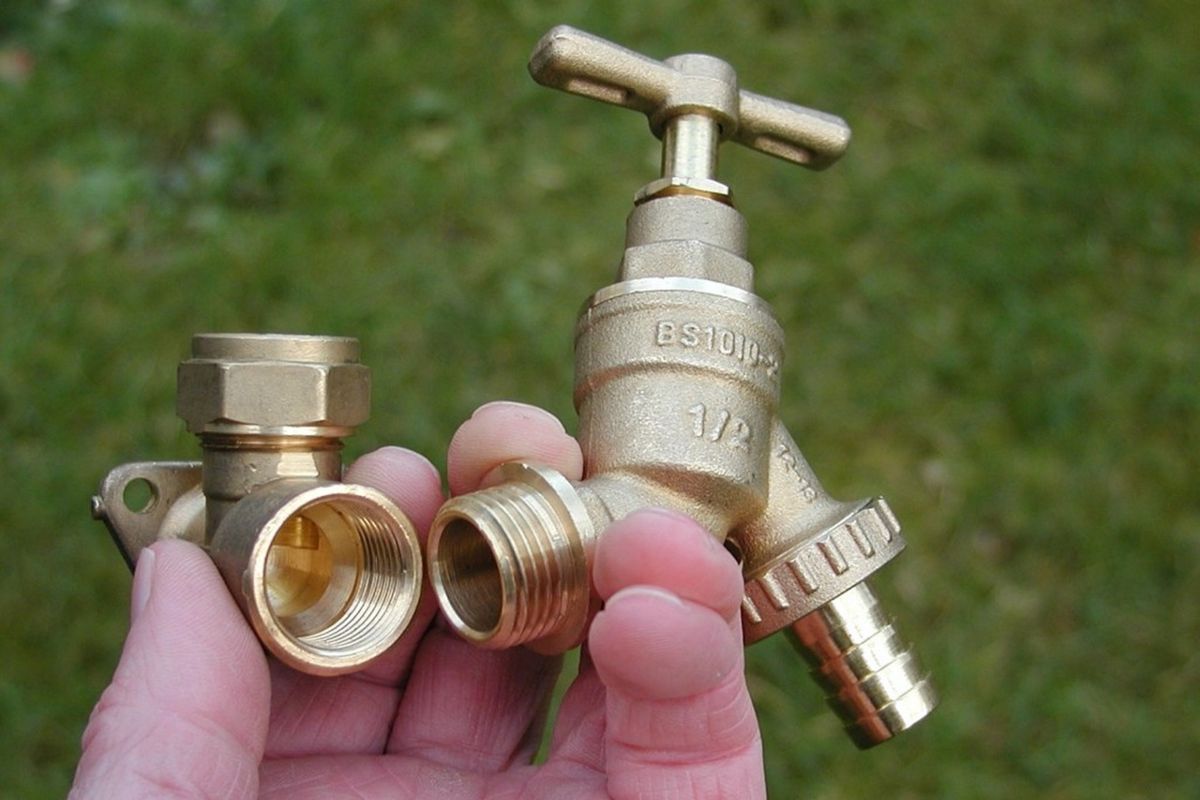 A hand holds two new brass plumbing fixtures, an L-shaped elbow joint and an outdoor water tap