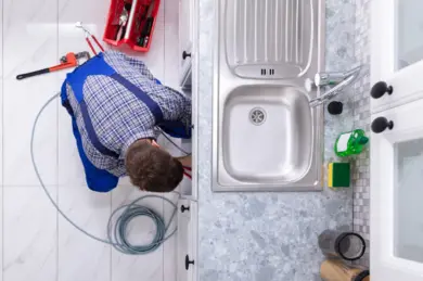 A plumber wearing blue overalls kneels on the floor beside a kitchen sink, working with tools and cables, with a coiled hose on the tiled floor and a red toolbox nearby.